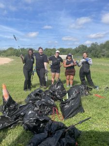 A group of volunteers including Sheridan Ray and Laisha Alvarado stand together outdoors during an environmental restoration event at Onion Creek Metropolitan Park. Trees and grassy parkland surround the group.